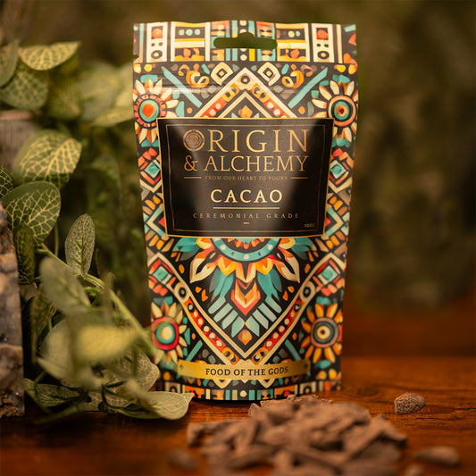 A package of 100g Origin & Alchemy Ceremonial Cacao with a colorful, patterned design displayed on a wooden surface with cacao beans scattered around.