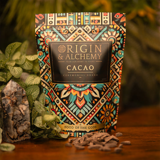 A 500g package of Origin & Alchemy Ceremonial Cacao with a colorful, patterned design, displaying the product name and a tagline 'Food of the Gods'. The package is presented on a wooden surface with a crystal and loose cacao pieces in the background.